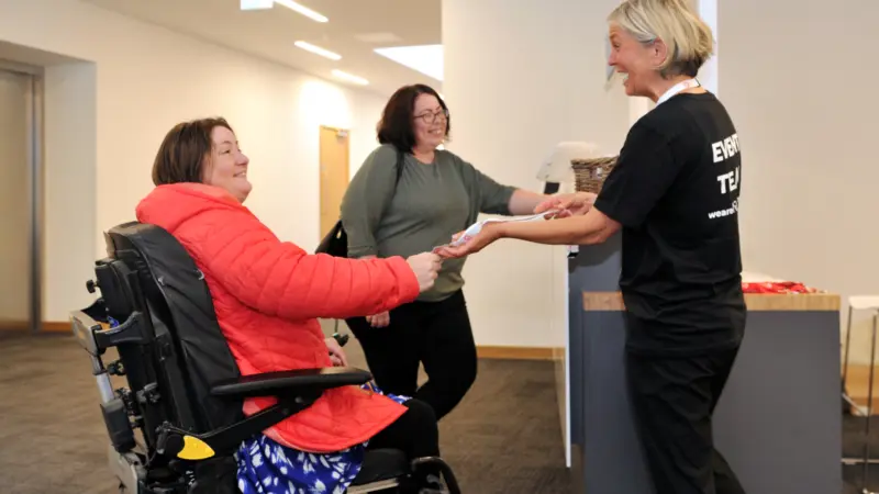 This image shows two people arriving for a Q event, one of the people is a wheelchair user. A member of staff is welcoming them to the event and handing over a lanyard, everyone is smiling.
